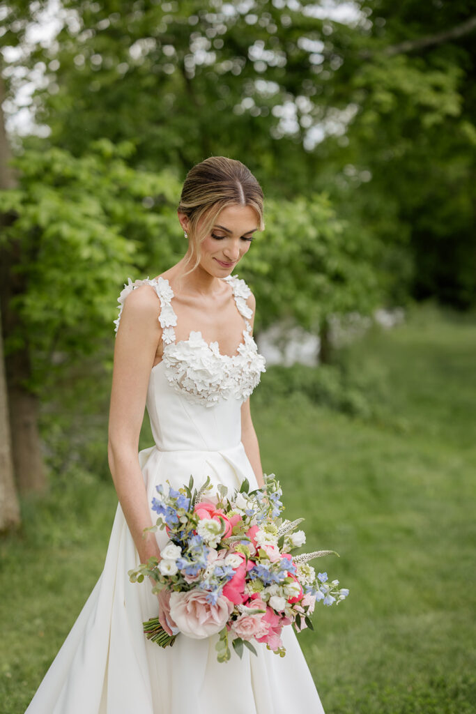The bride is looking down at her pink blue and white bouquet while standing in the grass shot my the timeless wedding photographer Monika Eisenbart