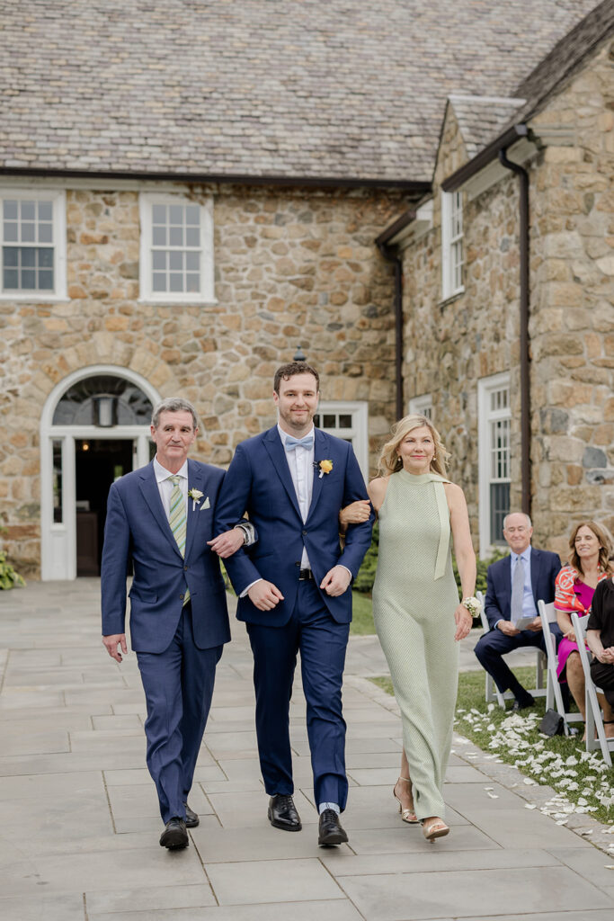 The groom in a blue walking down the aisle linking arms with his parents in blue and green, shot my the timeless wedding photographer Monika Eisenbart