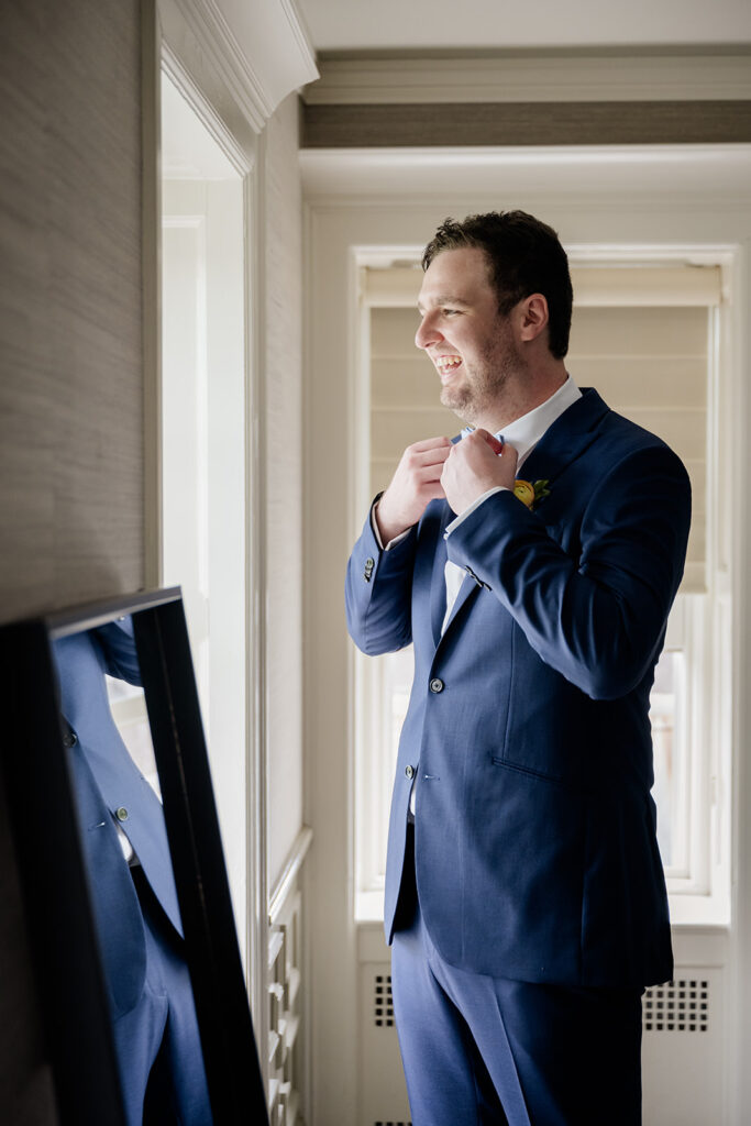 Groom in a blue suit adjusting his bowtie looking out the windown shot by the timeless wedding photographer Monika Eisenbart
