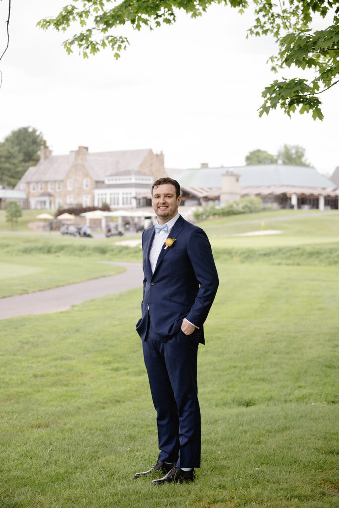 Groom standing in blue suit looking at the camera smiling while out in a field shot by the timeless wedding photographer Monika Eisenbart