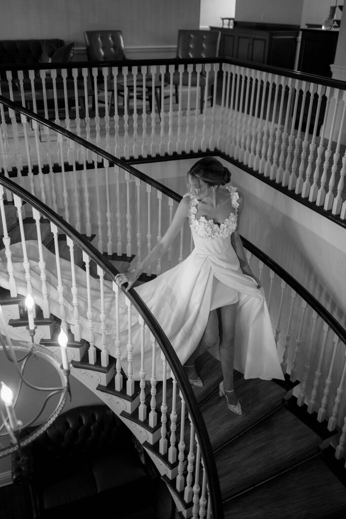 Black and white photo of a bride walking down a spiral staircase looking back over her shoulder shot by the timeless wedding photographer Monika Eisenbart