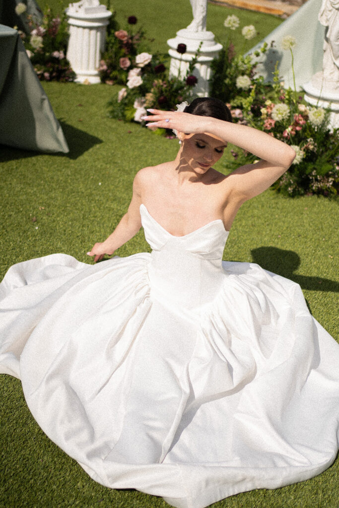 bride shielding her face from the sun while sitting on green grass in her wedding dress